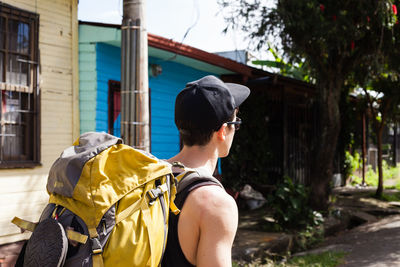 Side view of young man with backpack walking on road against house