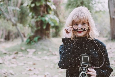 Portrait of young woman wearing sunglasses while holding vintage camera in park