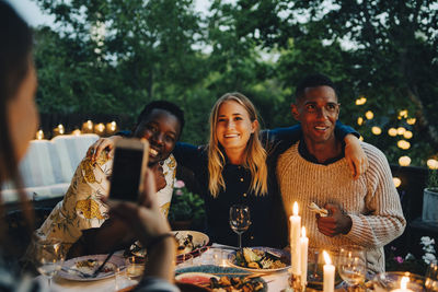 Woman photographing happy male and female friends sitting at table during dinner party