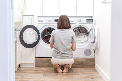 Rear view of woman sitting in front of washing machine at home