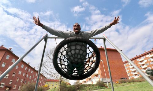 Low angle view of man with arms outstretched screaming on swing against sky
