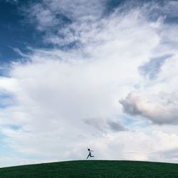 Low angle view of person jumping on grassy hill against cloudy sky