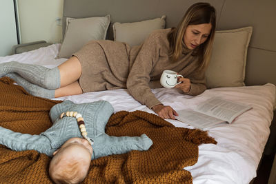 Mother holding coffee cup reading book while lying on bed with sleeping baby