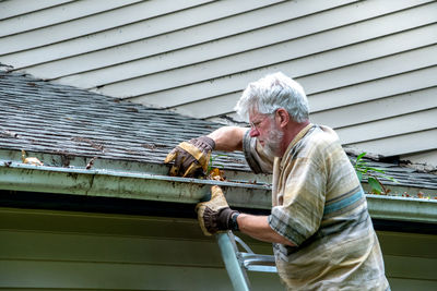 An older man stands on a ladder as he cleans out leaves and debris in the gutters on his home