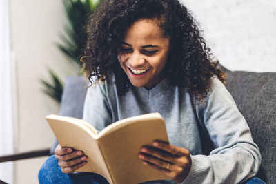 Smiling young woman reading book while sitting on seat