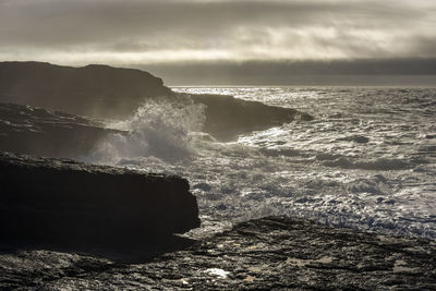 Scenic view of waves splashing on rocks at sea against sky