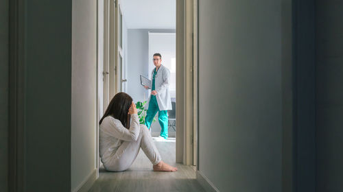 Doctor looking at female patient with mental disorder sitting on floor in hospital