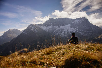 Man sitting on grassy field against mountains during sunny day