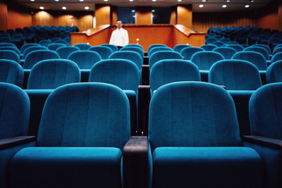Defocused mid adult man standing amidst blue empty chairs in auditorium