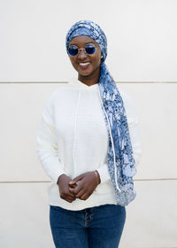 Self assured young african american female millennial in trendy sunglasses and traditional headscarf smiling and looking at camera against white wall