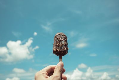 Cropped image of hand holding chocolate ice cream bar against blue sky