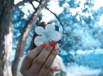 Woman holding artificial flower over face against trees