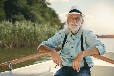 Portrait of senior man sitting in rowing boat on a lake wearing suspenders and summer hat