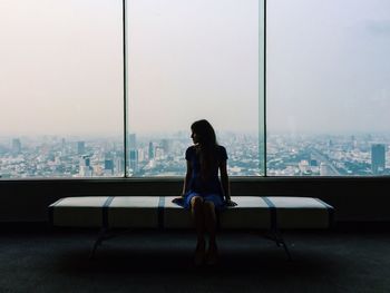 Young woman in blue dress sitting on seat against glass window in city