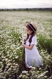 Portrait girl child in a white dress stands on a camomile field in a hat. bouquet of flowers