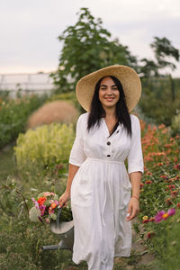 Beautiful brunette girl in a white dress and hat with watering can watering flowers