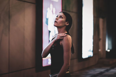 Young woman looking away while standing against wall at night