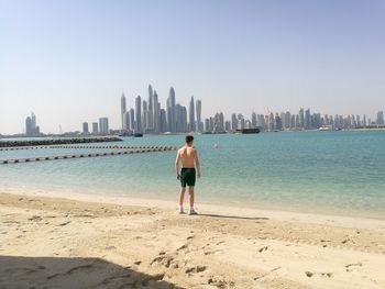 Rear view of shirtless man standing on shore at beach in city against clear sky