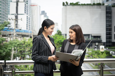 Smiling businesswomen looking at folder while standing against buildings in city