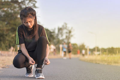 Full length of young woman tying shoelace while crouching on road against sky during sunset