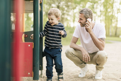 Smiling father talking on phone while looking at son playing in playground