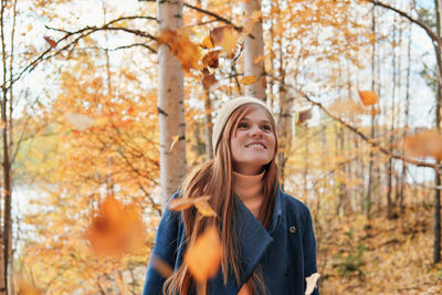 Happy young red haired woman smiling and looking at falling leaves and a tree.  autumn forest. 