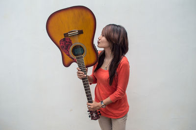 Young woman kissing guitar against white wall