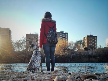 Rear view of pet owner with dog standing on by river against clear sky