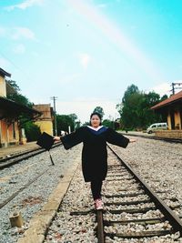 Young woman in graduation gown walking on railroad track against sky