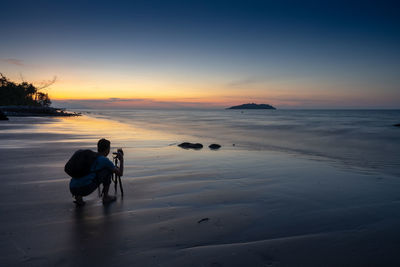 Rear view of man photographing sea at beach against sky during sunset