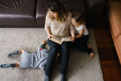 From above of young woman reading interesting fairytale in book while sitting on floor with little son and daughter at home