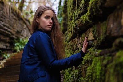 Side view portrait of young woman standing by mossy wall