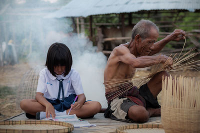 Schoolgirl studying while sitting with grandfather making wicker baskets