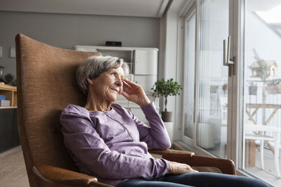 Portrait of senior woman sitting on armchair at home looking through window