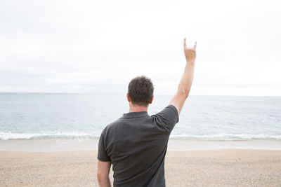 Rear view of man showing rock sign while standing at beach