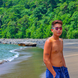 Portrait of shirtless young man wearing sunglasses standing on beach against trees