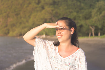 Smiling young woman shielding eyes while looking away at beach during sunny day