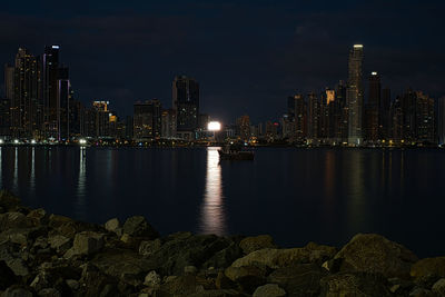 Illuminated buildings by river against sky at night