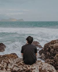 Rear view of woman sitting on rock by sea against sky