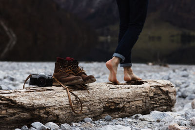 Side view of crop anonymous female traveler walking on weathered wooden log with boots and photo camera placed nearby on rocky waterfront in autumn day