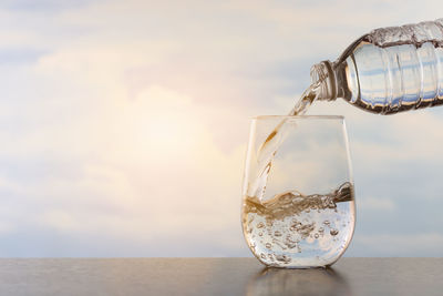 Close-up of water pouring from bottle in glass on table against sky