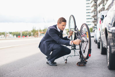 Businessman fixing bicycle chain on city street against sky