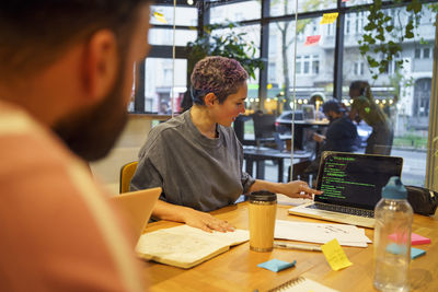 Non-binary it professional discussing coding on laptop to colleagues during meeting in office