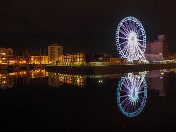 Illuminated ferris wheel by river against sky in city at night
