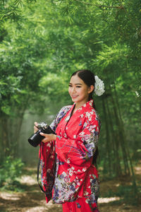 Smiling young woman wearing kimono holding digital camera in forest