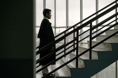 Full length serious man in graduation gown standing on stairs in building