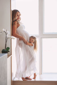 Beautiful pregnant mom with child near window at home. girl hugging belly and listening to baby