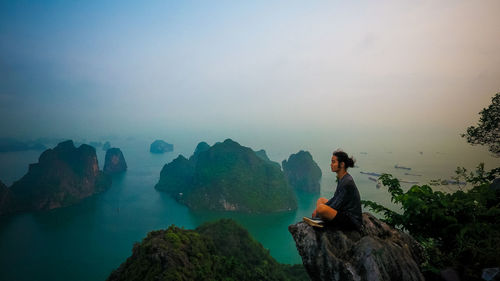 Young man sitting on rock against sky