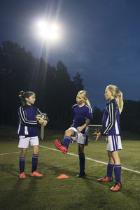 Girls talking while standing on soccer field against sky at night