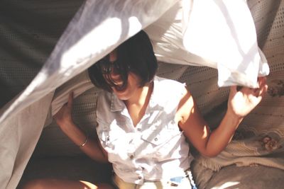 Young woman covering with sheet while sitting on bed with sunlight at home